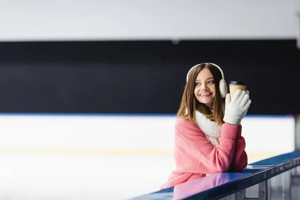 Mulher feliz em camisola de malha, abafadores de orelha e roupa de inverno segurando copo de papel na pista de gelo — Fotografia de Stock