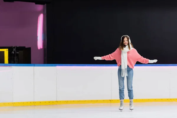 Piena lunghezza di donna allegra in manicotti orecchio bianco e sciarpa a maglia pattinaggio con le mani tese sulla pista di pattinaggio su ghiaccio — Foto stock