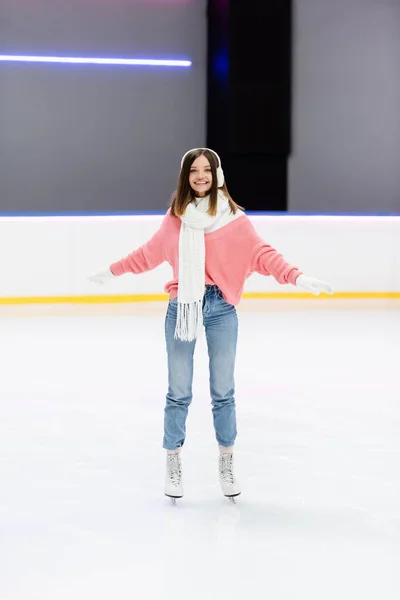 Full length of cheerful woman in white ear muffs and scarf skating with outstretched hands on ice rink — Stock Photo