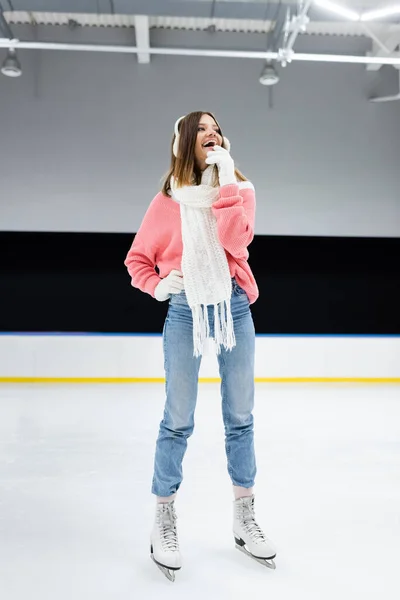 Full length of cheerful woman in white ear muffs and knitted scarf skating on ice rink — Stock Photo