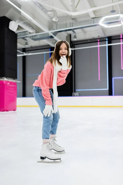Comprimento total de mulher alegre em regalos de orelha branca e patinação de cachecol de malha enquanto ri na pista de gelo — Fotografia de Stock