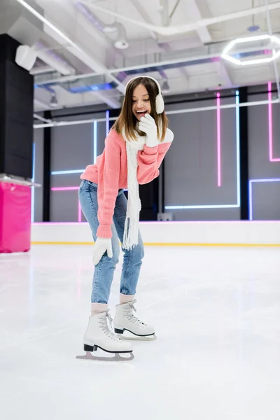 Longitud completa de feliz joven mujer en orejeras y bufanda patinaje mientras se ríe en pista de hielo - foto de stock