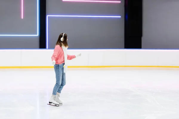 Longueur pleine de jeune femme en pull tricoté, cache-oreilles et foulard patinant sur patinoire — Photo de stock