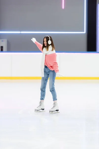 Full length of smiling young woman in ear muffs and scarf skating with outstretched hands on ice rink — Stock Photo