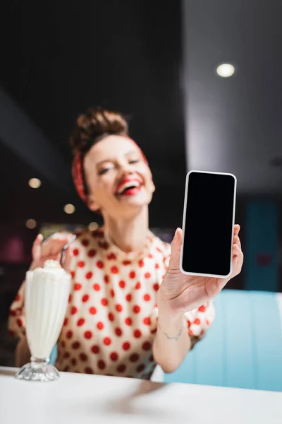 Mujer pin up borrosa y feliz sosteniendo teléfono inteligente con pantalla en blanco - foto de stock