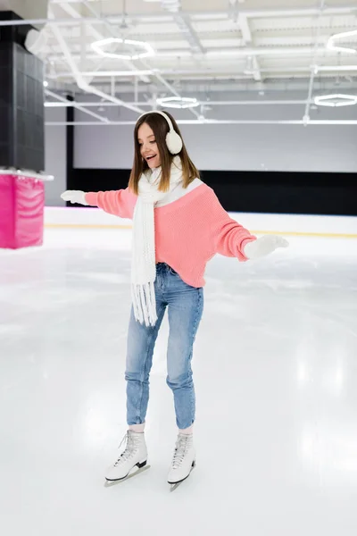 Feliz joven mujer en orejeras blancas y tejido bufanda patinaje con las manos extendidas en pista de hielo - foto de stock