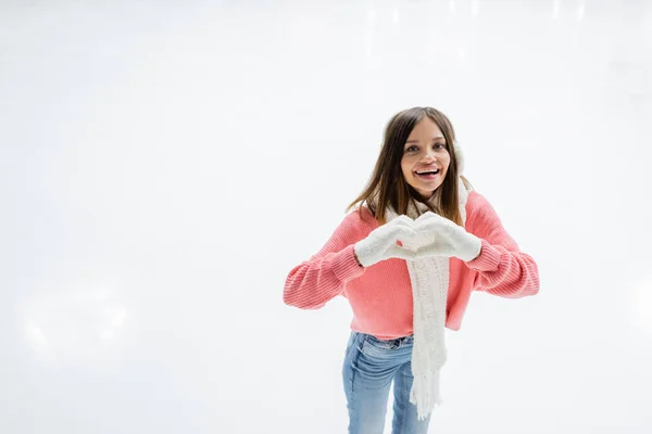Mujer joven feliz en orejeras blancas y bufanda de punto que muestra el signo del corazón en la pista de hielo - foto de stock
