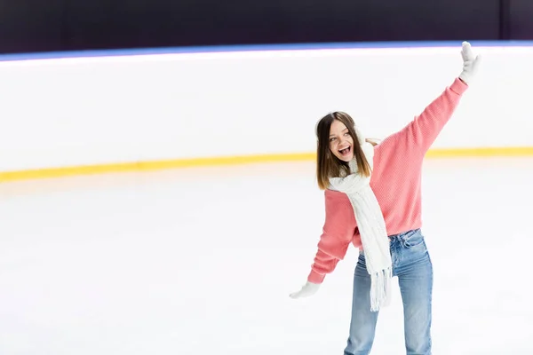 Amazed young woman in white ear muffs and knitted scarf skating with outstretched hands on ice rink — Stock Photo