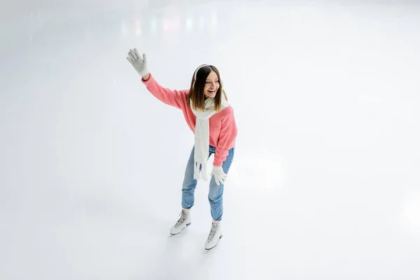 Vista ad alto angolo di stupito giovane donna in manicotti orecchio bianco e maglia pattinaggio maglione con mano tesa sulla pista di pattinaggio su ghiaccio — Foto stock