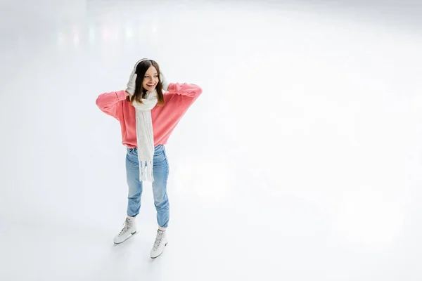 Vue grand angle de la jeune femme étonnée tenant des cache-oreilles blancs et debout sur la patinoire — Photo de stock
