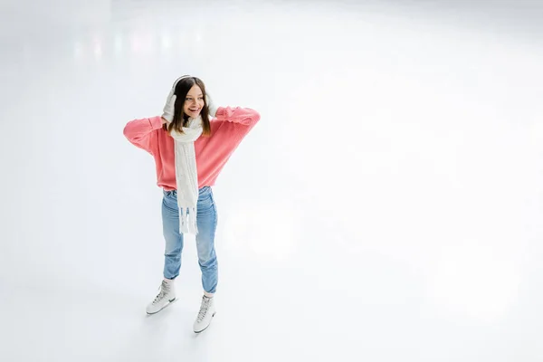 Vista de ángulo alto de la mujer joven asombrada sosteniendo orejeras blancas y de pie en pista de hielo - foto de stock