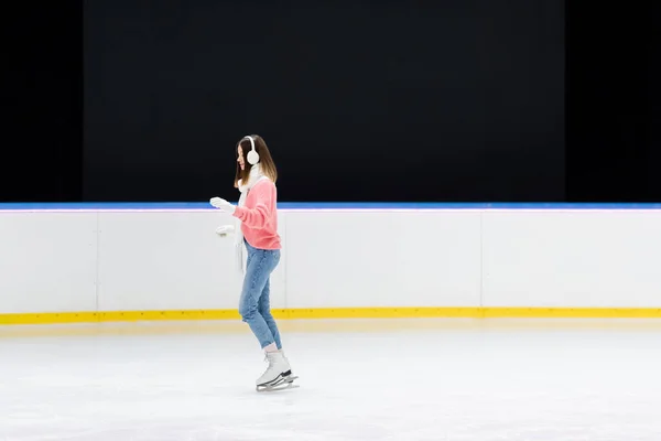 Vue latérale de la jeune femme en cache-oreilles blancs patinant sur la patinoire — Photo de stock