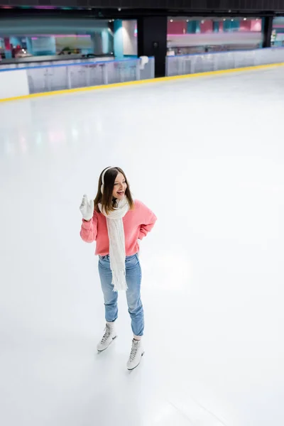 Full length of happy woman posing with hand on hip and waving hand on ice rink — Stock Photo