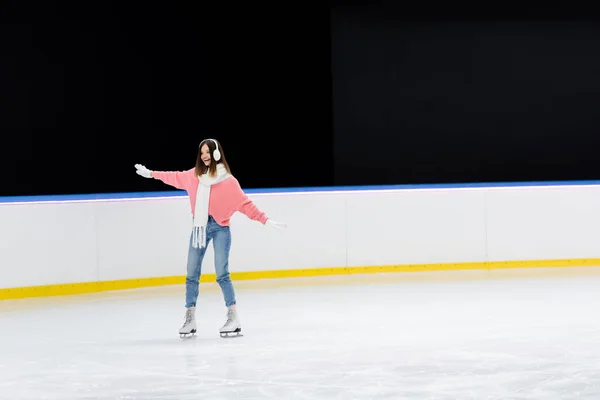 Longitud completa de la joven excitada en orejeras y bufanda patinaje con las manos extendidas en pista de hielo - foto de stock