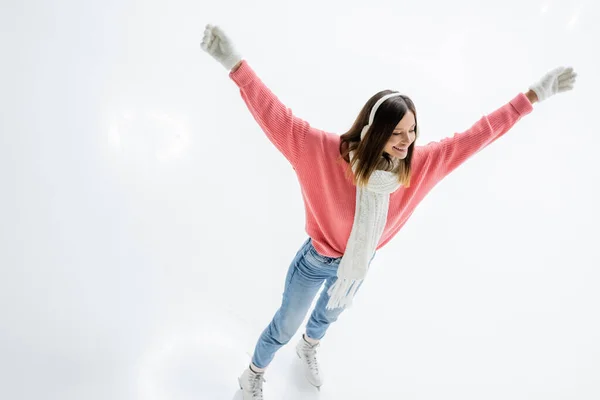 High angle view of of excited young woman in ear muffs and jeans skating with outstretched hands on ice rink — Stock Photo