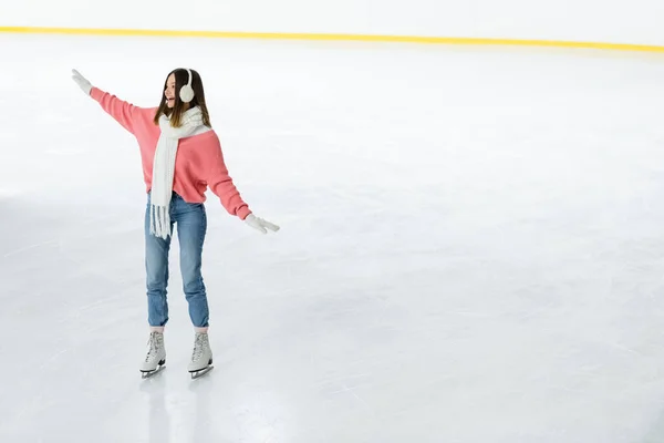 Volle Länge der staunenden jungen Frau in Ohrenschützer und Schal Schlittschuhlaufen mit ausgestreckten Händen auf der Eisbahn — Stockfoto