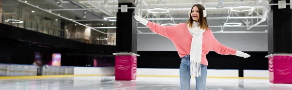 Joyeuse jeune femme en cache-oreilles et foulard patinant avec les mains tendues sur la patinoire, bannière — Photo de stock