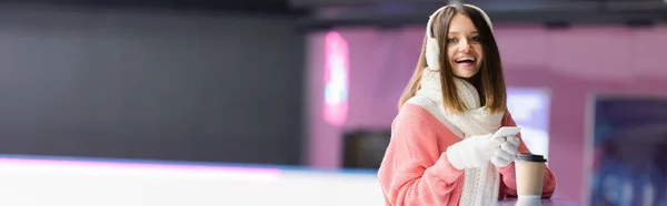 Mujer feliz en orejeras blancas y suéter rosa sosteniendo el teléfono celular cerca de la taza de papel, pancarta - foto de stock