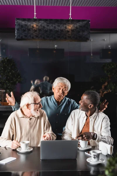 Изумленный азиат показывает вау-жест рядом со старшими межрасовыми друзьями, указывающими на ноутбук в кафе — стоковое фото