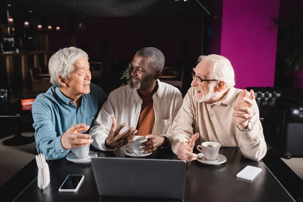 Feliz sénior amigos multiétnicos gesto mientras habla en la cafetería cerca de la computadora portátil y tazas de café — Stock Photo