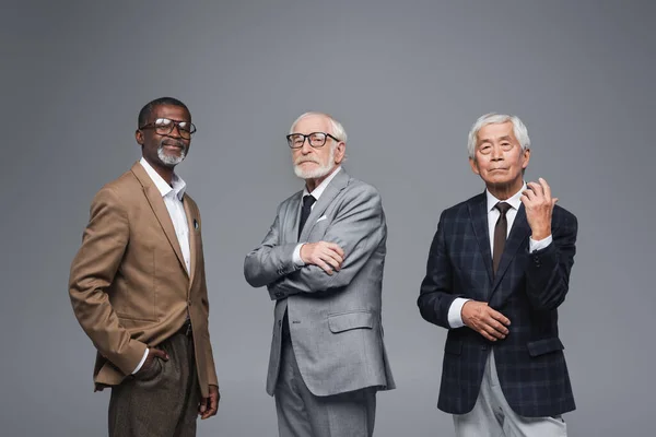 Socios de negocios multiétnicos de edad avanzada en ropa formal mirando a la cámara mientras están aislados en gris - foto de stock