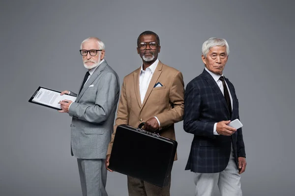 Senior multiethnische Geschäftsleute mit Vertrag, Aktentasche und Smartphone blicken isoliert auf graue Kamera — Stockfoto