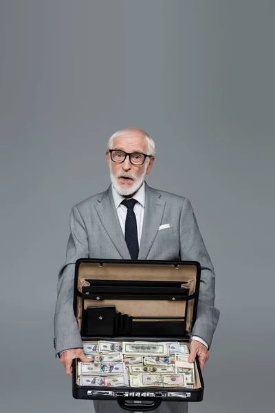 Amazed senior businessman showing briefcase full of dollars isolated on grey — Stock Photo