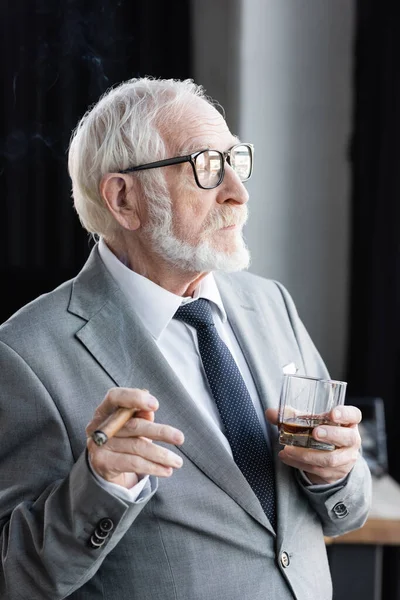 Elderly businessman with cigar and glass of whiskey looking away in office — Stock Photo