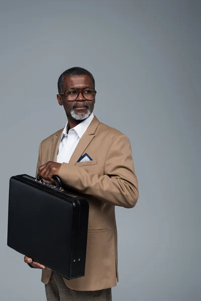 Anziano e fiducioso uomo d'affari afroamericano distogliendo lo sguardo mentre tiene la valigetta isolata su grigio — Foto stock