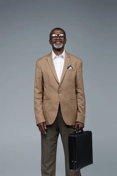 Positiver älterer afrikanisch-amerikanischer Geschäftsmann mit Aktentasche, der isoliert auf grau in die Kamera lächelt — Stockfoto