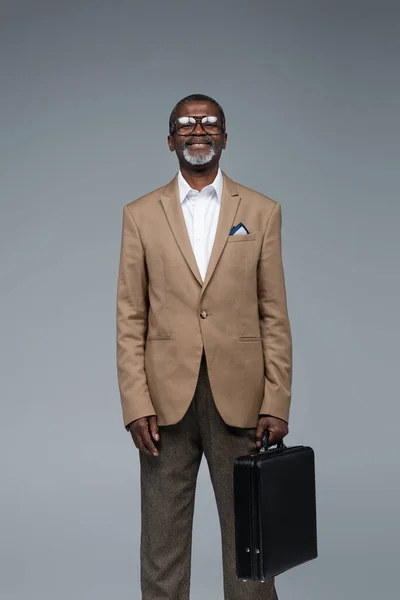 Positive senior african american businessman with briefcase smiling at camera isolated on grey — Stock Photo