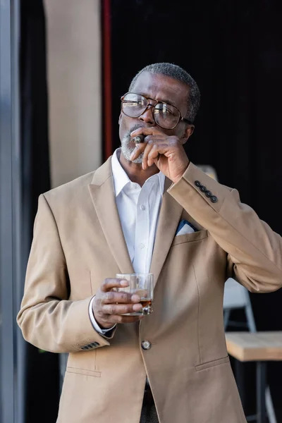 Empresário americano africano sênior fumar charuto enquanto segurando um copo de uísque — Fotografia de Stock