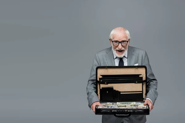 Verwunderter Senior-Geschäftsmann blickt in die Kamera, während er Aktentasche mit Geld in grau isoliert hält — Stockfoto