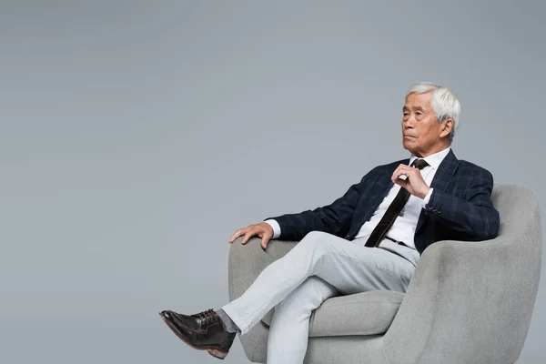 Senior y confidente asiático hombre de negocios mirando hacia otro lado mientras sentado en sillón aislado en gris - foto de stock