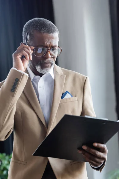 Nachdenklicher afrikanisch-amerikanischer Geschäftsmann justiert Brille, während er im Büro auf Klemmbrett schaut — Stockfoto