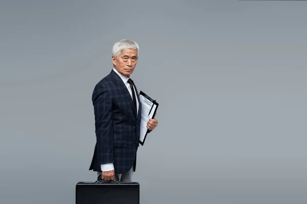Älterer und selbstbewusster asiatischer Geschäftsmann mit Aktentasche und Vertrag, der isoliert auf grau schaut — Stockfoto