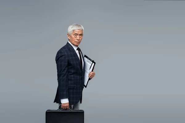Elderly and confident asian businessman with briefcase and contract looking at camera isolated on grey — Stock Photo