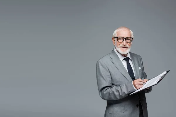 Smiling senior businessman looking at camera while signing contract isolated on grey — Stock Photo