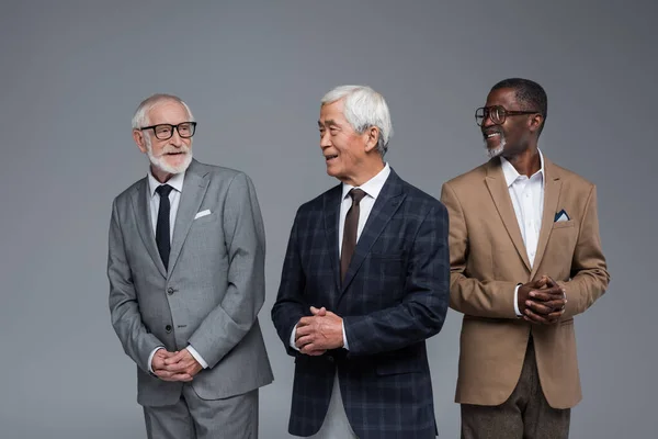 Idosos empresários multiétnicos em desgaste formal sorrindo uns para os outros isolados em cinza — Fotografia de Stock