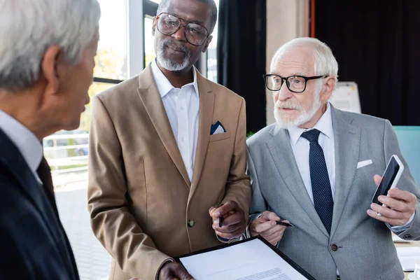 Hombres de negocios multiétnicos senior mostrando contrato y teléfono inteligente con pantalla en blanco a colega borrosa - foto de stock