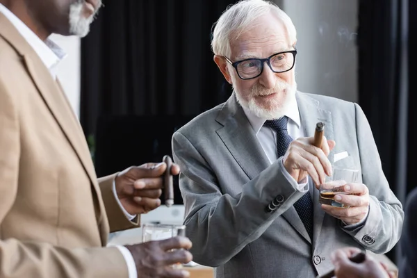 Hombre de negocios senior con cigarro y whisky mirando hacia otro lado durante la discusión con un borroso colega afroamericano - foto de stock