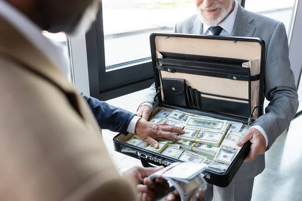 Partial view of senior businessman holding briefcase with money near blurred interracial business partners — Stock Photo