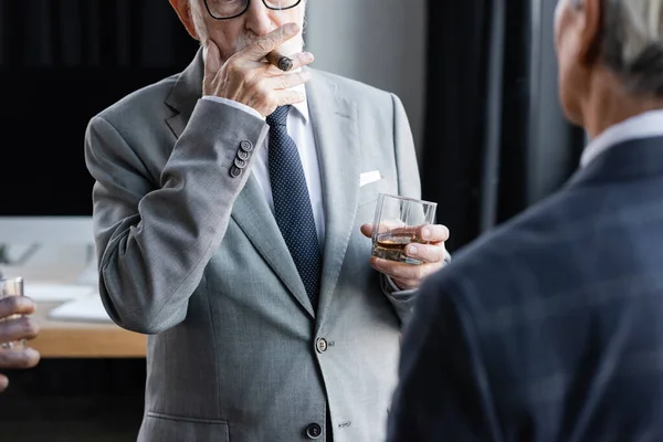 Partial view of senior businessman with glass of whiskey smoking cigar near blurred interracial business partners — Stock Photo