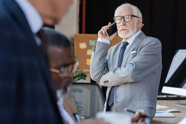 Пожилой бизнесмен в очках, разговаривающий по мобильному телефону рядом с размытыми многонациональными коллегами — стоковое фото