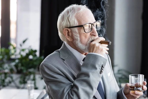 Senior businessman with glass of whiskey looking away while smoking cigar in office — Stock Photo