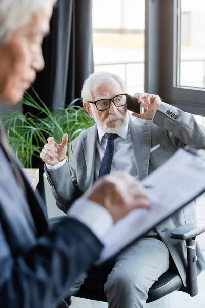 Senior businessman hablando en smartphone cerca borrosa asiático colega con portapapeles - foto de stock