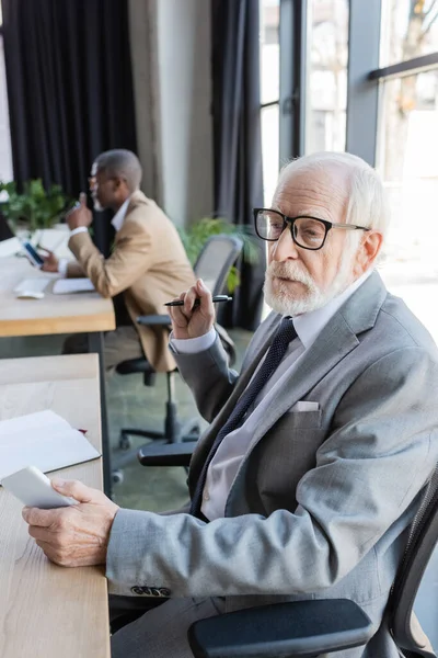 Anziano uomo d'affari in occhiali in possesso di telefono cellulare mentre collega afro-americano che lavora su sfondo sfocato — Foto stock