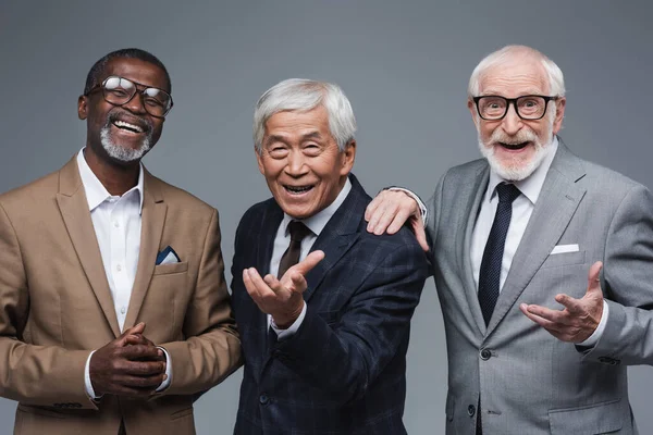 Senior multiethnic businessmen with outstretched hands near smiling african american colleague isolated on grey — Stock Photo