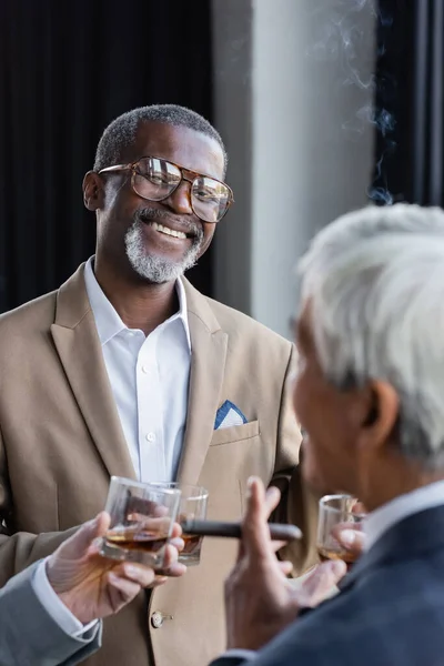 Cheerful african american businessman in eyeglasses standing with cigar and glass of whiskey near blurred senior colleagues — Stock Photo