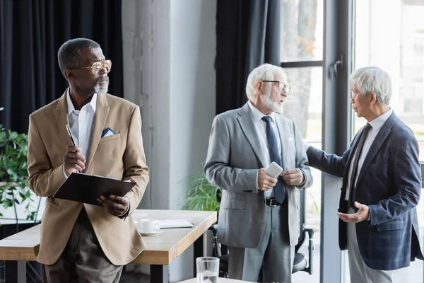 Senior africano americano hombre de negocios de pie con portapapeles cerca de hablar interracial colegas — Stock Photo